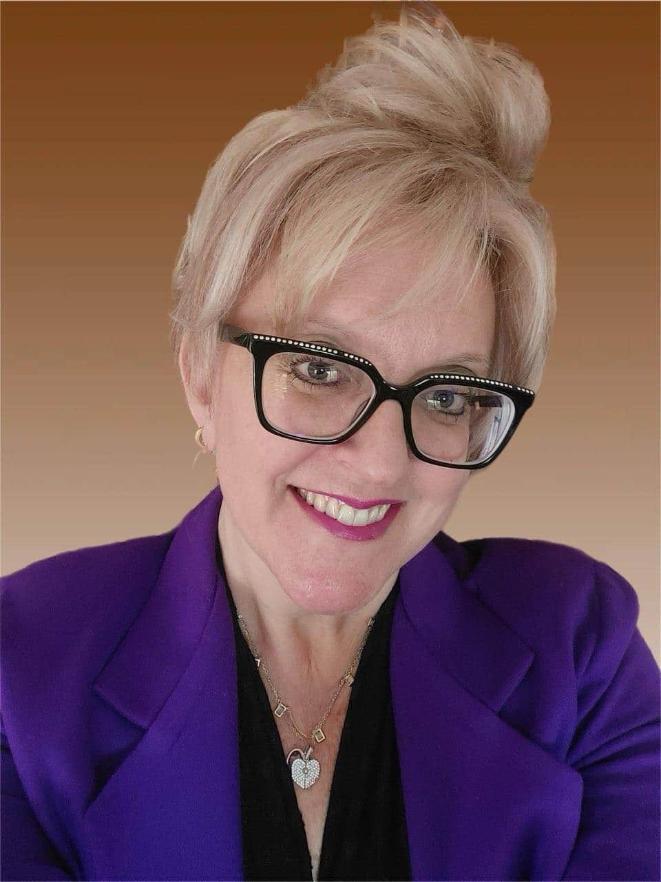 A blonde woman with her hair in a bun is smiling. She wears a purple suit coat and black glasses.